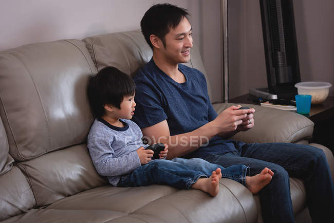Вид сбоку счастливых азиатских отца и сына, играющих вместе в видеоигры, сидя дома на диване — стоковое фото