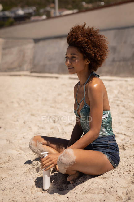 Vue latérale de jeune jolie femme mixte se détendre sur la plage tout en tenant lotion solaire dans sa main par une journée ensoleillée
. — Photo de stock