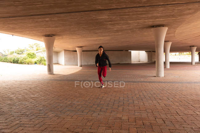 Vista frontal da jovem mulher de raça mista correndo sob uma ponte na cidade — Fotografia de Stock