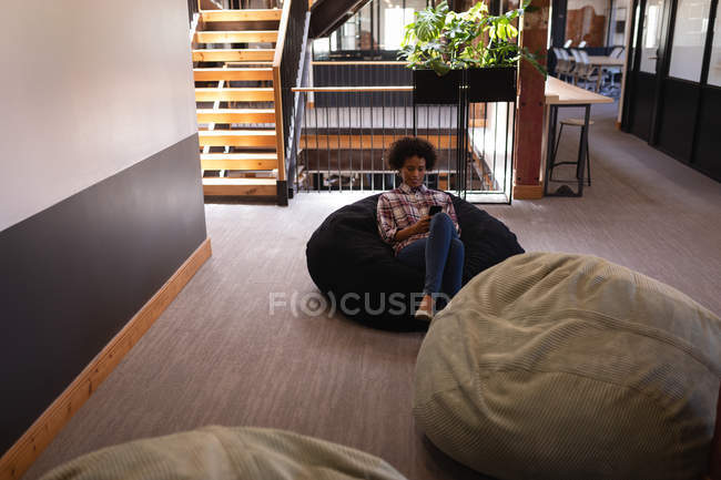 Вид спереду красива бізнес-леді змішаної раси, що використовує мобільний телефон, розслабляючись у мішку з квасолею в сучасному офісі — стокове фото