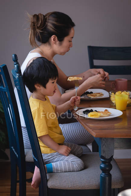 Vista lateral de uma mãe e filho asiáticos desfrutando de café da manhã na mesa de jantar na cozinha em casa — Fotografia de Stock