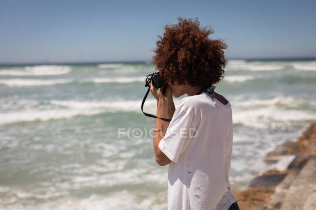 Vista lateral da jovem mestiça clicando fotos com câmera digital na praia em um dia ensolarado — Fotografia de Stock