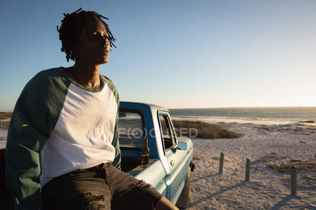 Seitenansicht eines jungen afrikanisch-amerikanischen Mannes, der an einem sonnigen Tag auf einem Auto am Strand sitzt — Stockfoto