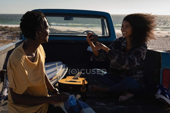 Vista lateral da mulher afro-americana capturando foto do homem afro-americano enquanto estava sentado no carro na praia no dia ensolarado — Fotografia de Stock