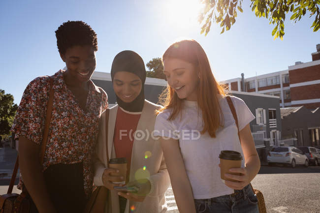 Вид спереду змішаної раси друзів-жінок, що використовують мобільний телефон, стоячи на вулиці в сонячний день — стокове фото