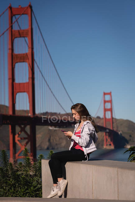 Вид збоку красивої жінки, використовуючи мобільний телефон, сидячи на стіні — стокове фото