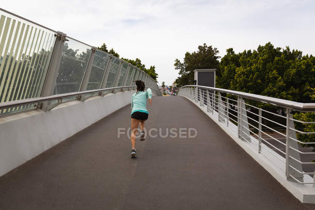 Rückansicht einer jungen Mischlingsfrau, die auf einer Fußgängerbrücke in der Stadt läuft — Stockfoto