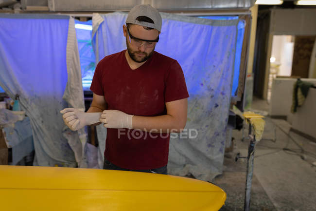 Front view of handsome Caucasian putting latex gloves on while standing in front of surfboard in workshop. — Stock Photo