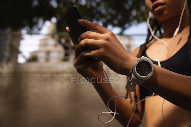 Seção intermediária da mulher de raça mista usando telefone celular enquanto ouve música em fones de ouvido na cidade — Fotografia de Stock