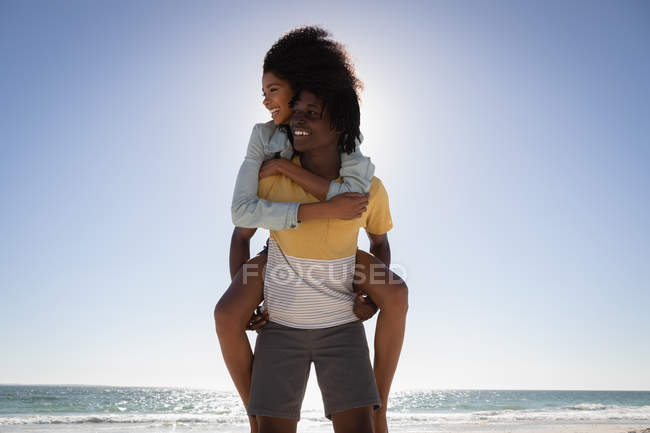 Front view of young African American man giving young African American woman piggyback at beach on a sunny day — стокове фото
