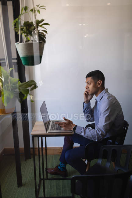 Вид збоку молодого змішаної раси бізнесмен розмовляє на мобільному телефоні, використовуючи ноутбук в сучасному офісі з рослинами перед собою — стокове фото