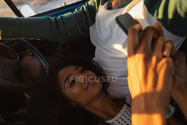 Vista de ángulo alto del joven afroamericano que conduce mientras que la mujer de raza mixta bastante usando el teléfono móvil en el coche - foto de stock