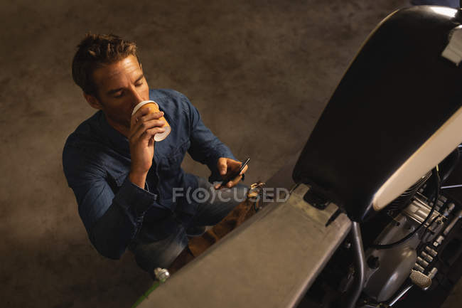 Hochwinkelaufnahme eines kaukasischen männlichen Fahrradmechanikers, der Kaffee trinkt, während er sein Handy in der Garage benutzt — Stockfoto