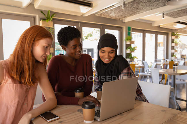 Vista lateral de jovens mestiços amigos do sexo feminino interagindo uns com os outros ao usar laptop em um café — Fotografia de Stock