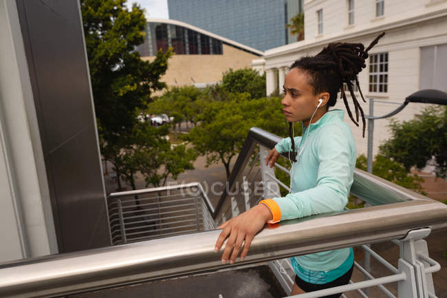 Vista lateral da jovem mulher de raça mista ouvindo música em fones de ouvido nas escadas da cidade — Fotografia de Stock