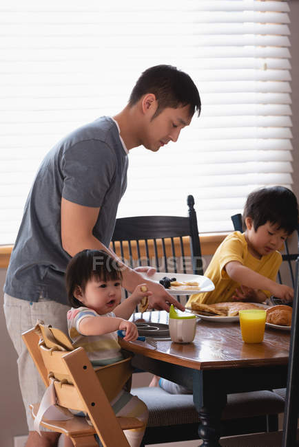 Vista laterale di un padre asiatico che serve la colazione ai suoi figli a tavola in cucina a casa — Foto stock