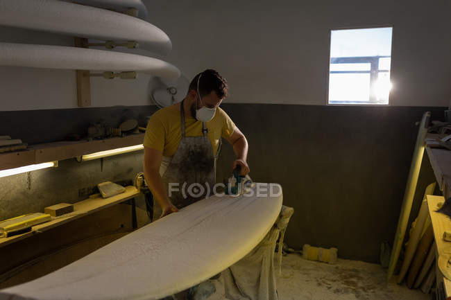 Vista frontal do homem caucasiano moldando a prancha de surf com máquina enquanto usava uma máscara protetora em uma oficina — Fotografia de Stock
