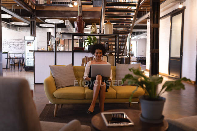 Vista frontale di bella donna d'affari mista seduta sul divano giallo mentre sta usando il computer portatile contro gli arredi — Foto stock