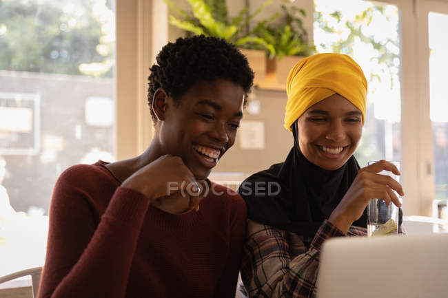 Side view of young African American female friends interacting with each other while using laptop in restaurant — Stock Photo