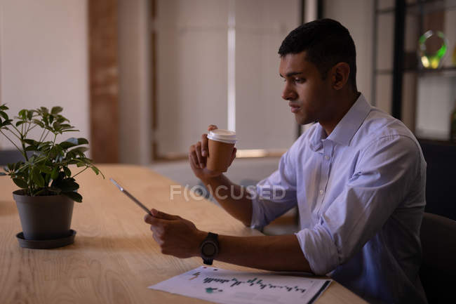 Seitenansicht eines hübschen jungen gemischten Geschäftsmannes, der am Schreibtisch im modernen Büro ein digitales Tablet benutzt, während er eine Kaffeetasse in der Hand hält — Stockfoto