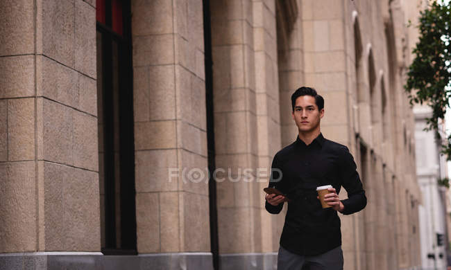 Vista frontal del hombre asiático guapo de pie en la calle mientras sostiene el teléfono móvil y el café en la mano - foto de stock