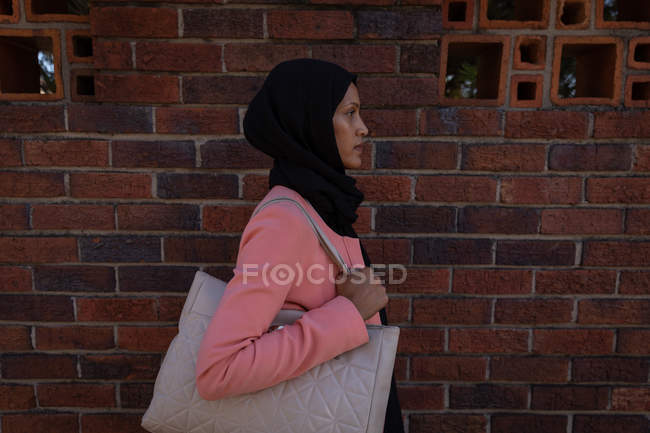 Вид збоку вдумлива серйозна змішана жінка, що йде на тротуарі поруч з деякий час тримаючи сумку — стокове фото