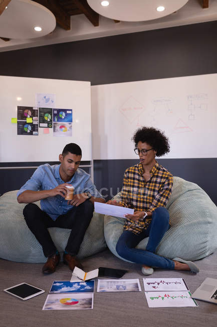 Вид спереду молодих бізнесменів змішаної раси, які обговорюють графік, сидячи в сучасному офісі — стокове фото