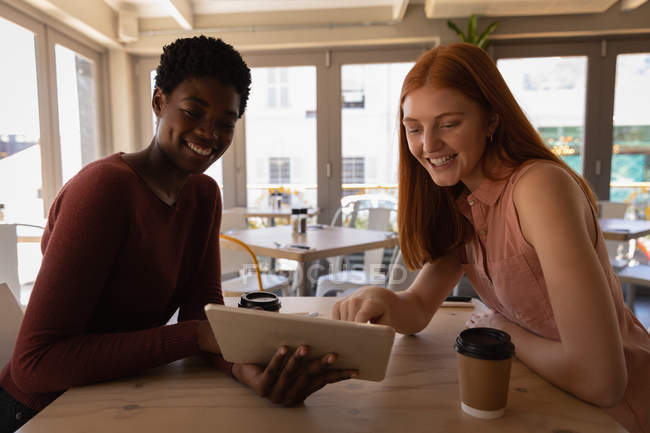 Vista lateral de jóvenes amigas de raza mixta usando tableta digital en un café - foto de stock