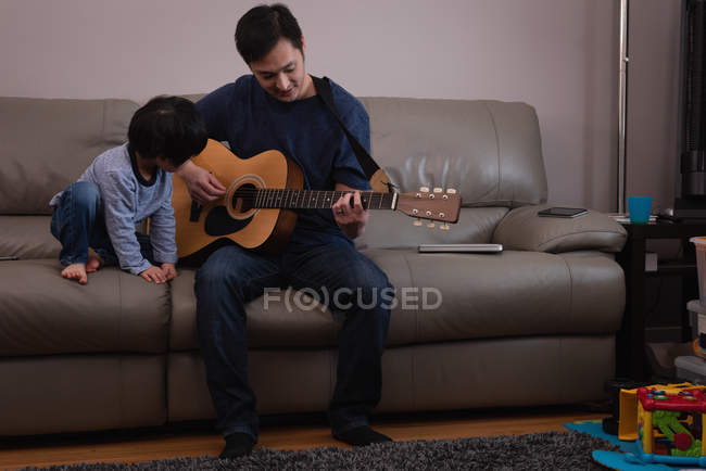 Vista frontal de um pai asiático tocando guitarra para seu filho no sofá em casa — Fotografia de Stock