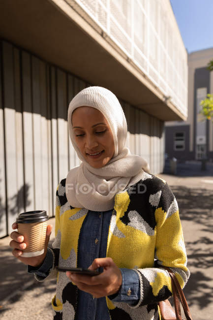 Вид спереду на молоду змішану жінку, яка використовує мобільний телефон, маючи каву на вулиці в сонячний день — стокове фото