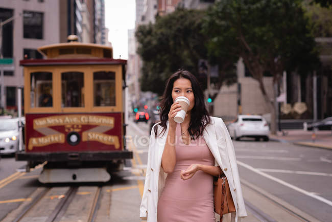 Vorderansicht einer nachdenklichen Asiatin, die auf der Straße vor einer Straßenbahn steht und Kaffee trinkt — Stockfoto