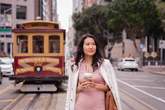 Vista frontal de la mujer asiática reflexiva de pie frente a un tranvía en la calle mientras toma café - foto de stock