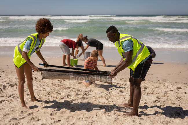 Vue latérale de jeunes volontaires multi-ethniques utilisant une passoire spéciale pour nettoyer la plage par une journée ensoleillée — Photo de stock