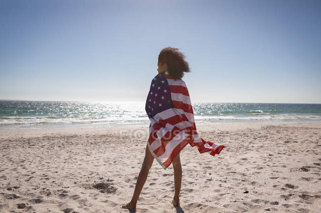 Vista trasera de la joven mujer envuelta bandera americana mientras está de pie en la playa en un día soleado - foto de stock