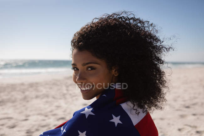Ritratto di giovane donna afroamericana con bandiera americana in piedi in spiaggia in una giornata di sole — Foto stock