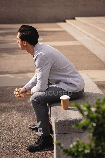 Вид збоку молодої азіатської людини, що має каву і сніданок, сидячи на кам'яних лавку — стокове фото