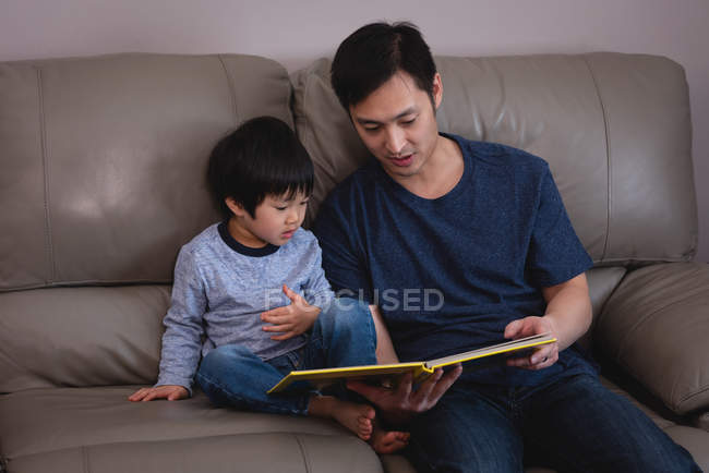 Vista frontale del padre asiatico che legge il libro di storie a suo figlio mentre è seduto sul divano a casa — Foto stock