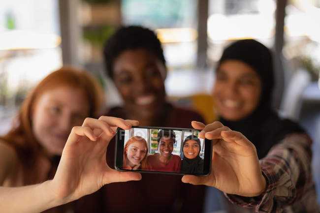 Vue de face de jeunes amies métisses prenant selfie avec téléphone portable dans un café — Photo de stock