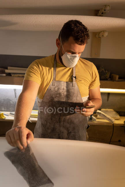 Vue de face de l'homme caucasien avec masque de protection de la bouche en utilisant un téléphone portable lors du nettoyage de la planche de surf dans un atelier . — Photo de stock
