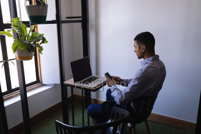 Vue latérale du jeune homme d'affaires métis utilisant un téléphone portable tout en travaillant sur un ordinateur portable au bureau — Photo de stock