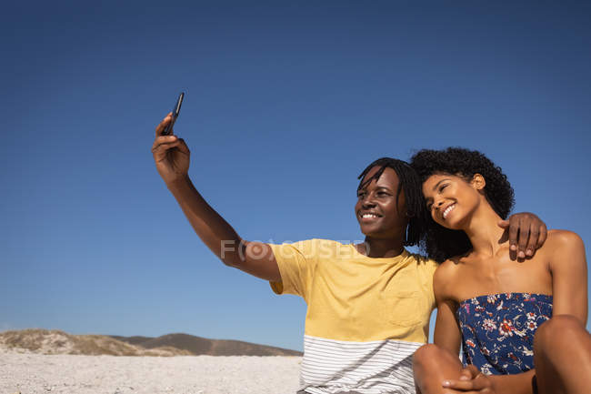 Vista frontal do jovem casal afro-americano feliz tomando selfie enquanto está sentado na praia em um dia ensolarado — Fotografia de Stock