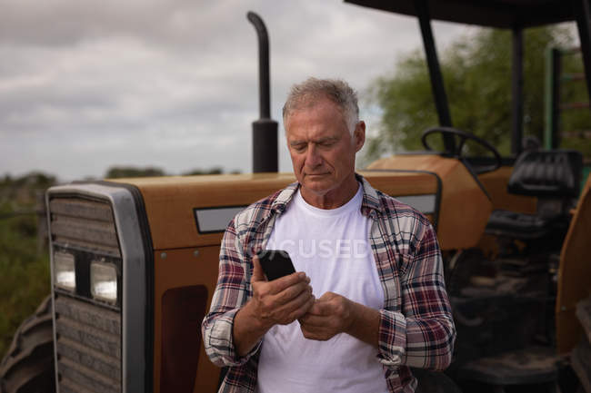 Вид на старшого Кавказького чоловічого фермера за допомогою мобільного телефону, стоячи перед його трактор в ферму — стокове фото