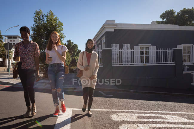 Вид спереди счастливых подруг смешанной расы, взаимодействующих друг с другом во время перехода дороги в солнечный день — стоковое фото