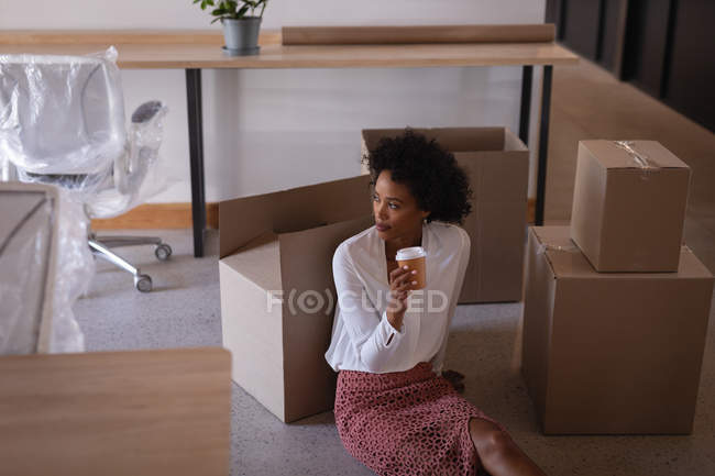 Vista de bajo ángulo de la pensativa mujer de negocios de raza mixta tomando café mientras está sentada en el suelo contra cajas de cartón en la oficina moderna - foto de stock