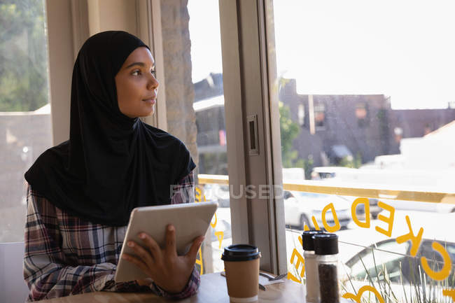 Vista frontal da jovem pensativa no hijab usando tablet digital em um café — Fotografia de Stock