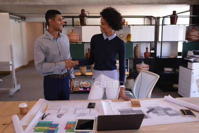 Front view of happy mixed-race business people shaking hands with plans in foreground on desk. — Stock Photo