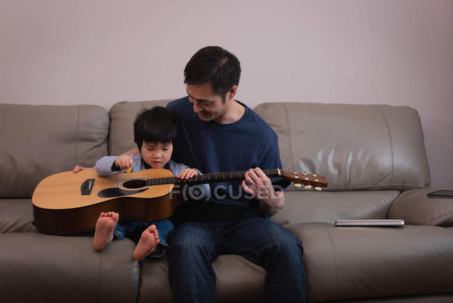 Vista frontal de um pai asiático feliz ensinando guitarra para seu filho no sofá em casa — Fotografia de Stock