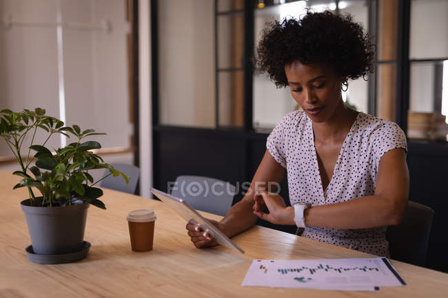 Vista lateral da bela jovem mulher de negócios de corrida mista verificando o tempo enquanto usa tablet digital sentado no escritório moderno com um café na mesa — Fotografia de Stock
