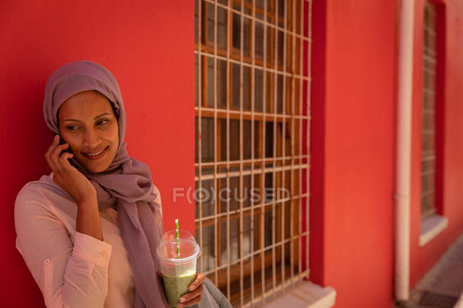 Вид сбоку на красивую расовую женщину, разговаривающую по мобильному телефону, прислонившуюся к стене и держащую смузи в солнечный день — стоковое фото