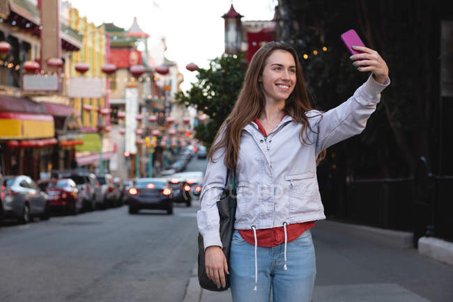 Vista frontal de la hermosa joven mujer caucásica tomando selfie mientras está de pie en paseo lateral en la ciudad - foto de stock